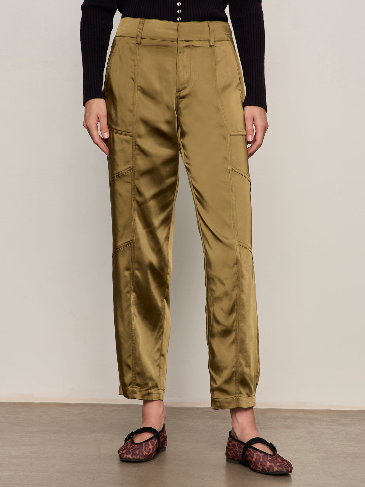 A person stands against a plain light wall wearing Sanctuary Clothing's sahara satin tapered pant in dirty martini, paired with a long-sleeve black top and brown leopard-print shoes. The photo is cropped at the shoulders.