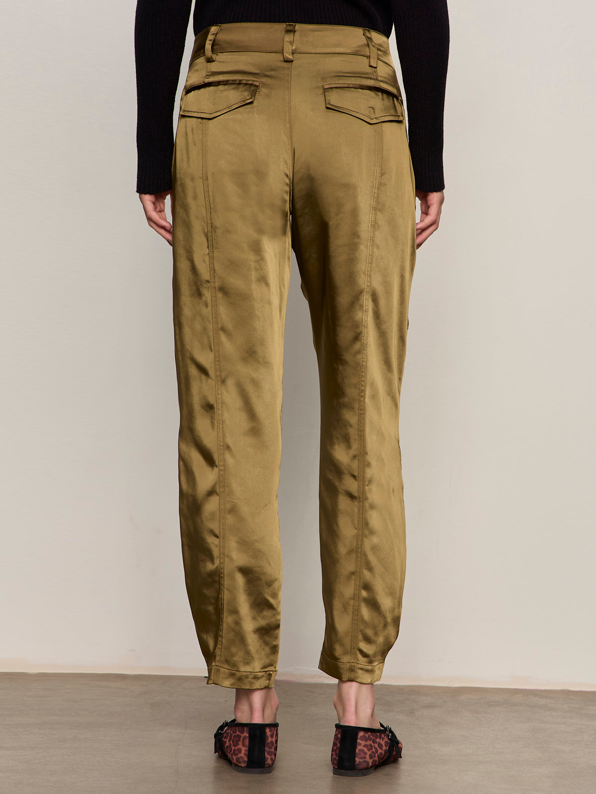 A person faces away, wearing Sanctuary Clothing’s sahara satin tapered pant in dirty martini, featuring back pockets, tapered legs, and paired with a black long-sleeve top and animal-print flats.