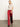A woman stands indoors against a plain light background, wearing Sanctuary Clothing's satin track pant in crimson with white side stripes, paired with a shiny beige short-sleeve top and red shoes.