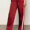 A person wears Sanctuary Clothing's satin track pant crimson—shiny red with white side stripes and a relaxed fit—paired with red heeled shoes. The photo, cropped at the torso, highlights the standout pants.