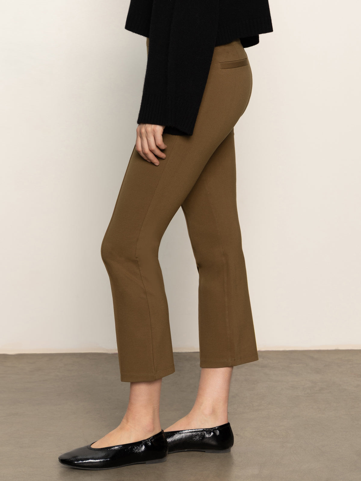 A person stands sideways on a gray floor, wearing Sanctuary Clothing's fairway legging in dirty martini, paired with a black long-sleeve top and black ballet flats against a plain, light background.