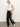 A woman stands against a plain background in Sanctuary Clothing’s coated twill track pant black, featuring white side stripes. She pairs them with a white sleeveless top and white sneakers with black accents, posing confidently with one hand in her pocket.