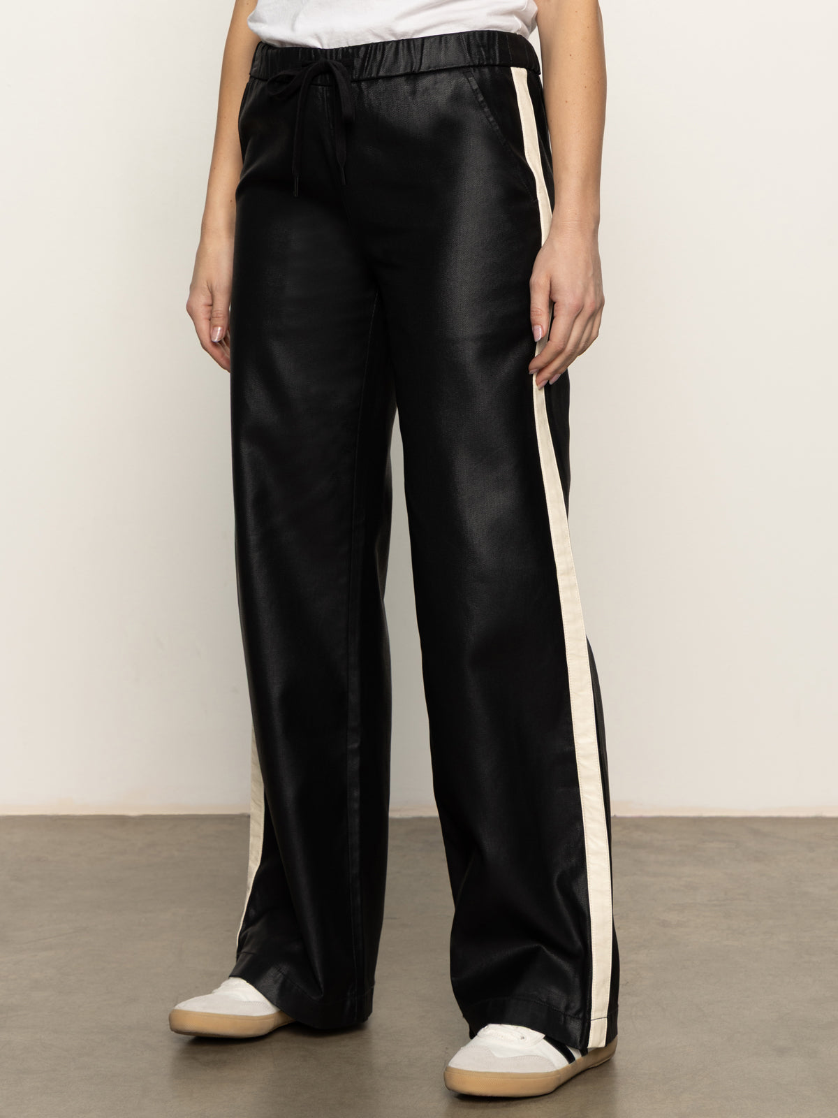 A person wears Sanctuary Clothing's coated twill track pant in black with white side stripes, a white T-shirt, and white sneakers, standing on a concrete floor against a plain light background.