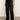 A person wears Sanctuary Clothing's coated twill track pant in black with white side stripes, a white T-shirt, and white sneakers, standing on a concrete floor against a plain light background.