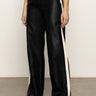 A person wears Sanctuary Clothing's coated twill track pant in black with white side stripes, a white T-shirt, and white sneakers, standing on a concrete floor against a plain light background.
