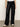 A person models the Sanctuary Clothing coated twill track pant in black, featuring wide legs and white side stripes, paired with white sneakers and a white top. Their head is cropped out against a neutral background.