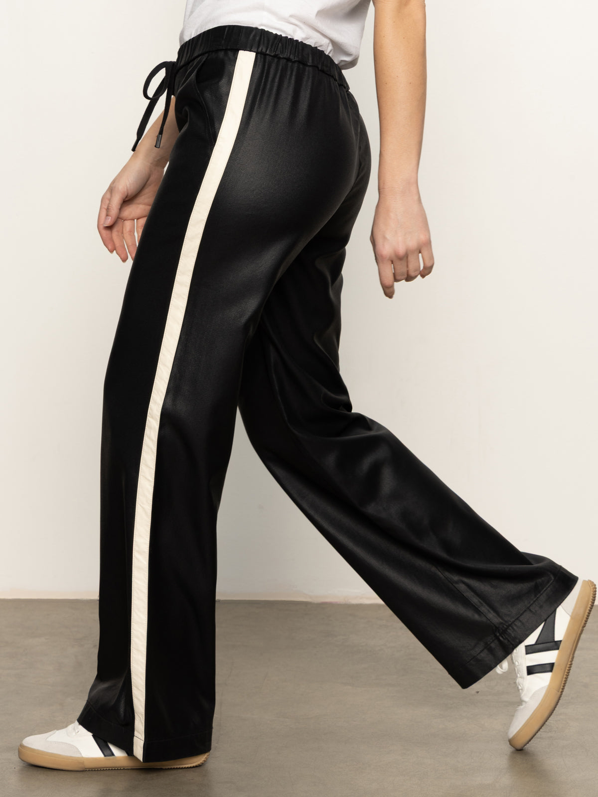 A person wearing Sanctuary Clothing's coated twill track pant in black, paired with a white shirt and white sneakers with black stripes, walking indoors on a gray floor.