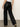 Someone wearing Sanctuary Clothing's coated twill track pant in black, featuring a white side stripe, paired with a white shirt and beige shoes, stands on a gray floor facing away from the camera.