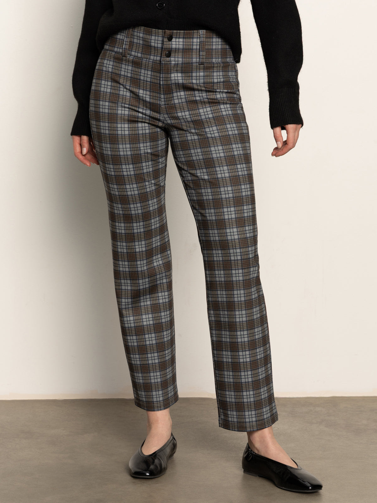 A person stands against a plain wall wearing Sanctuary Clothing’s 90s straight legging burnt olive plaid petites, paired with a black sweater and black ballet flats. Their head is out of frame.
