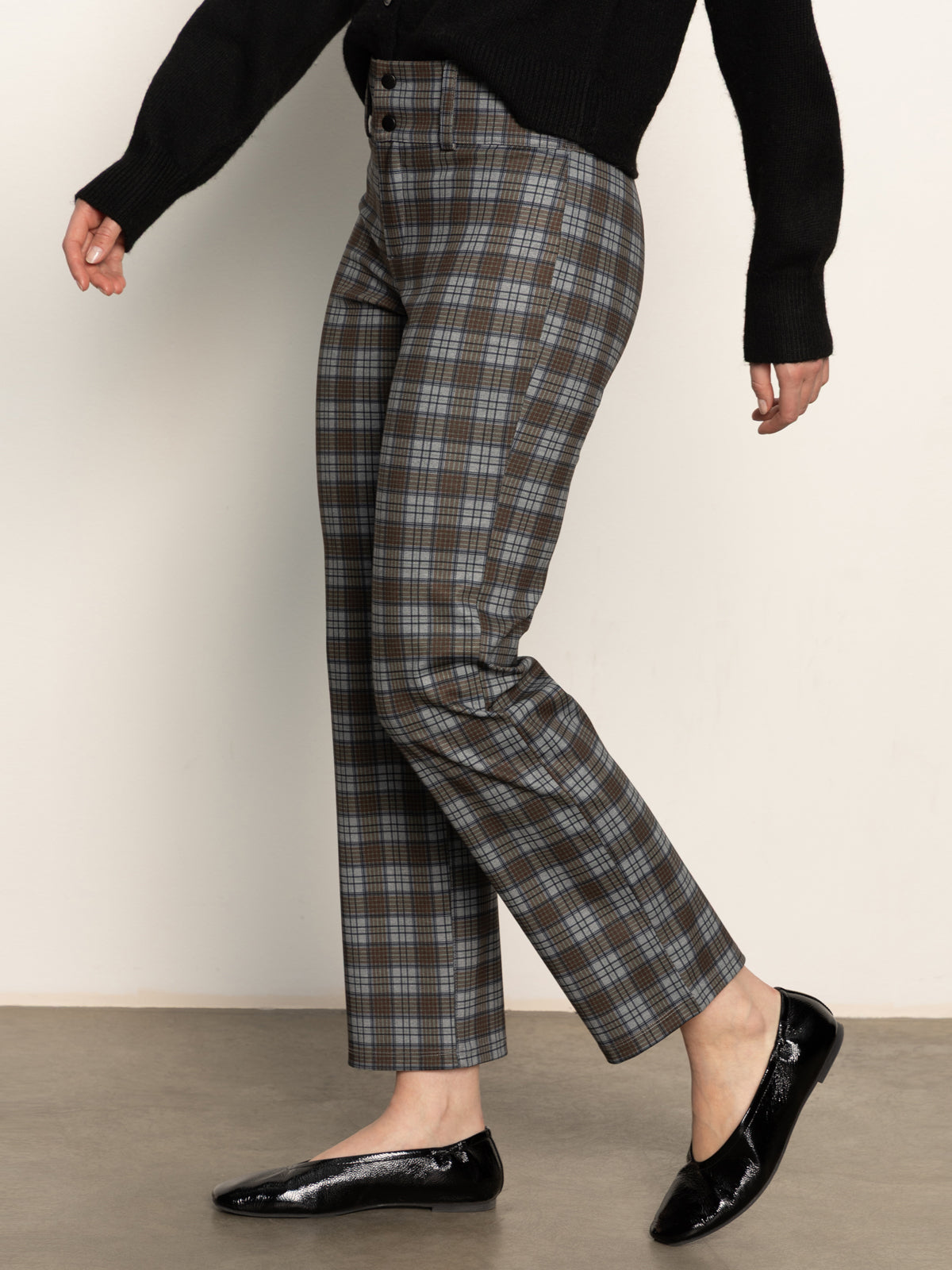 A person wearing Sanctuary Clothing’s 90s straight legging burnt olive plaid petites, a black long-sleeve sweater, and black patent leather flats stands against a plain light background. Their face is not visible.