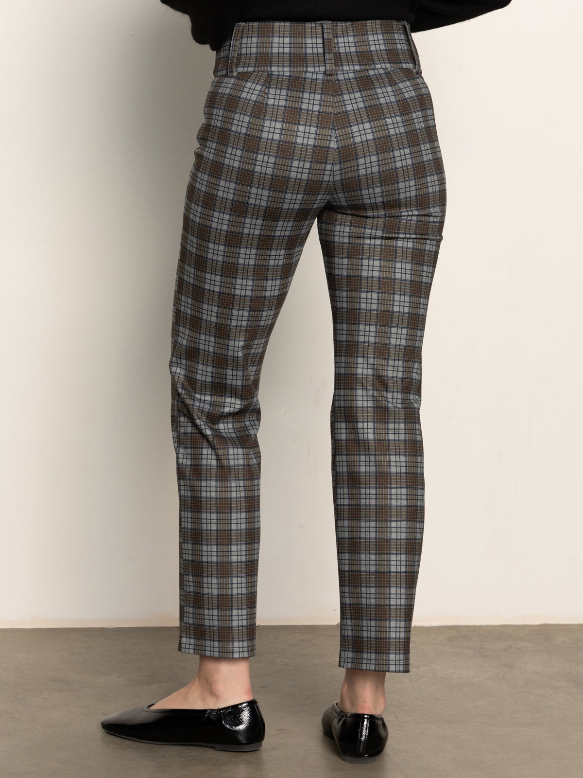 A person is seen from behind wearing Sanctuary Clothing's 90s straight legging burnt olive plaid petites, featuring a high-waisted, slim-fit design in brown, blue, and gray hues, paired with black flats against a plain backdrop.