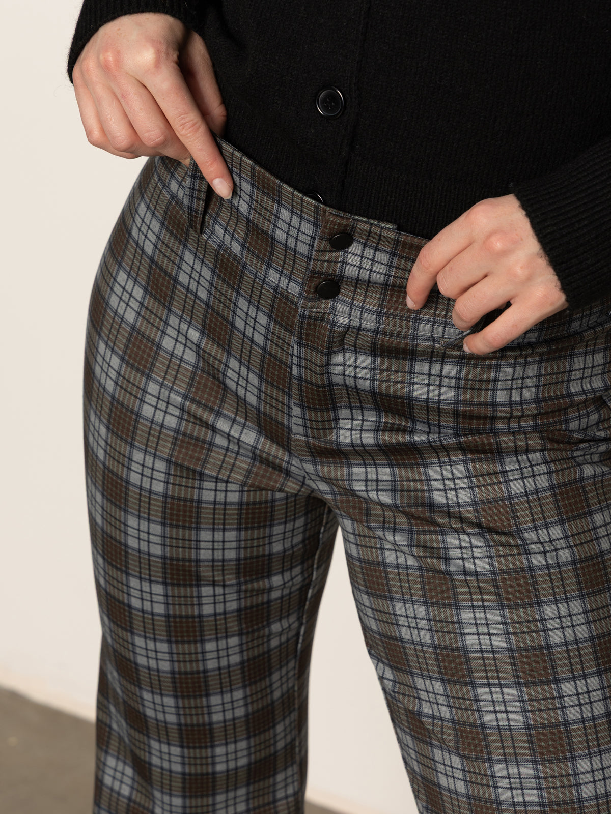 A person wearing a black sweater and pointing at the waistband of Sanctuary Clothing's 90s straight legging burnt olive plaid petites pants.