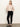 A woman stands indoors by a plain light wall, gazing to the side with hands behind her back. She wears black 90s straight legging by Sanctuary Clothing, a light pink buttoned cardigan, and black flat shoes.