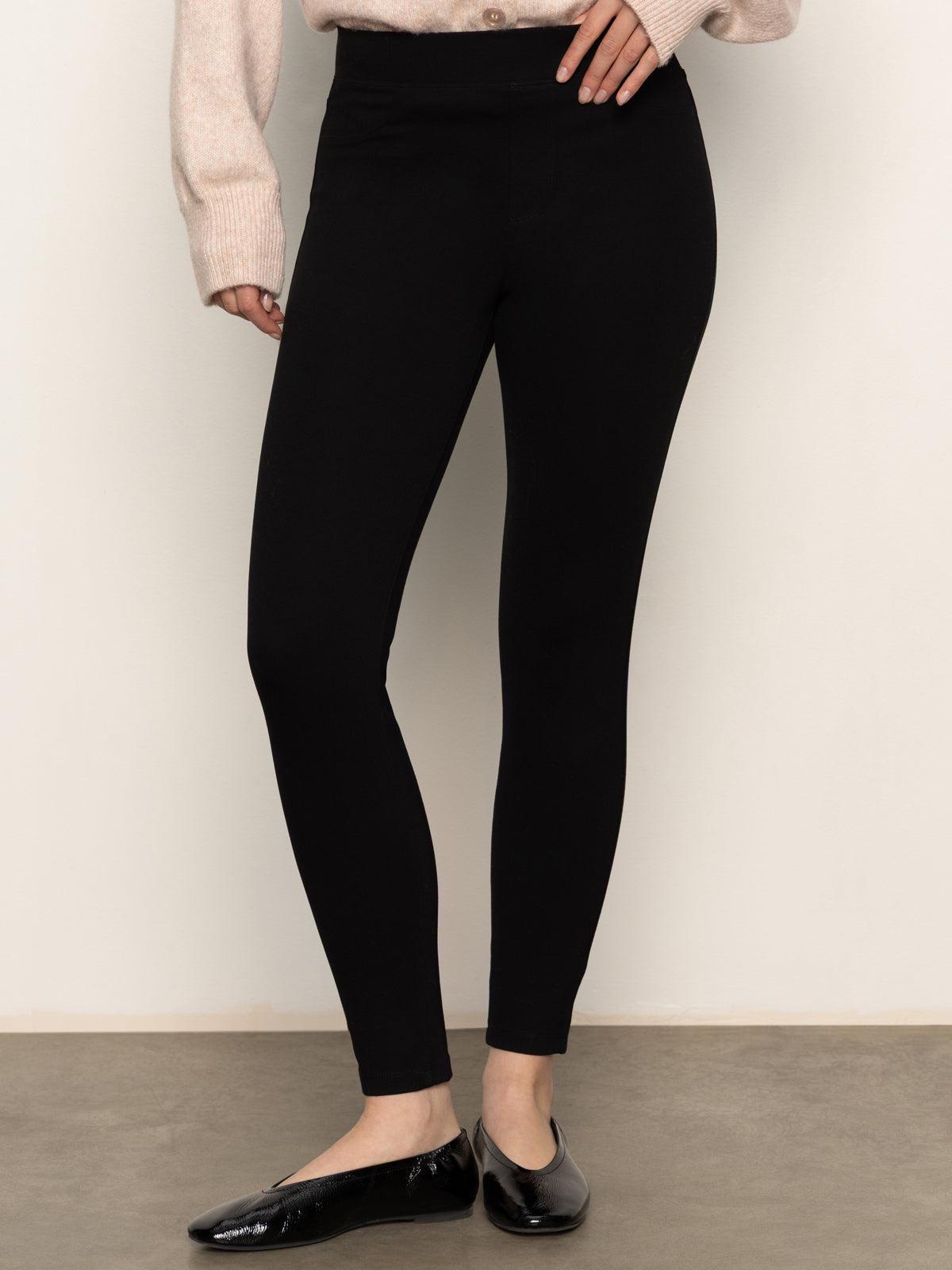 A person wears Sanctuary Clothing’s 90s Straight Legging in black, a light beige sweater, and black ballet flats, standing against a plain light wall with one hand on their hip.