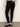 A person wears Sanctuary Clothing’s 90s Straight Legging in black, a light beige sweater, and black ballet flats, standing against a plain light wall with one hand on their hip.
