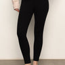 A person wears Sanctuary Clothing’s 90s Straight Legging in black, a light beige sweater, and black ballet flats, standing against a plain light wall with one hand on their hip.