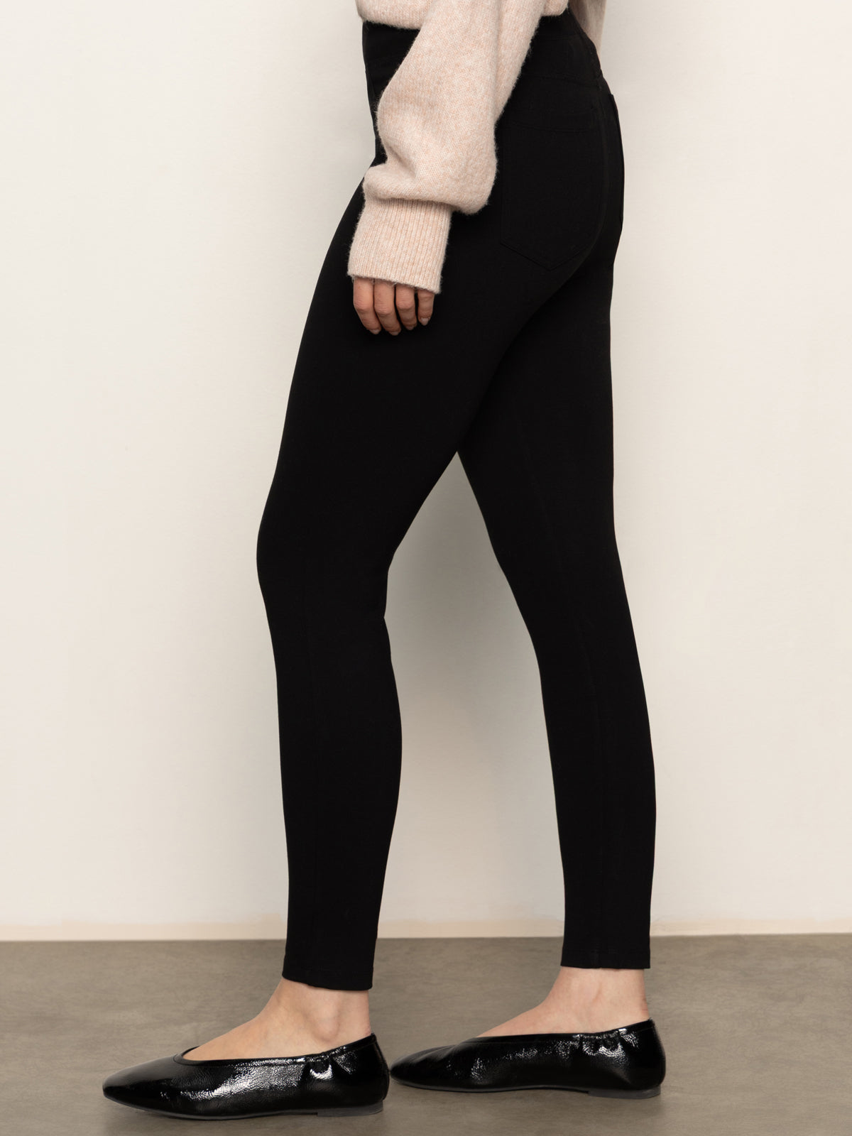 A woman stands sideways against a plain background, wearing the Sanctuary Clothing 90s straight legging black pants with a light long-sleeve sweater and black shiny ballet flats.