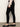 A woman stands sideways against a plain background, wearing the Sanctuary Clothing 90s straight legging black pants with a light long-sleeve sweater and black shiny ballet flats.