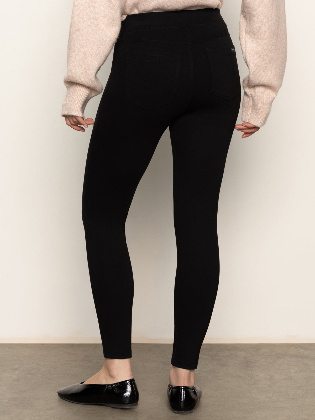 A person faces away, wearing Sanctuary Clothing’s 90s straight legging black, a light beige wrist-length sweater, and black shiny flats against a neutral background.