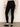 A person faces away, wearing Sanctuary Clothing’s 90s straight legging black, a light beige wrist-length sweater, and black shiny flats against a neutral background.