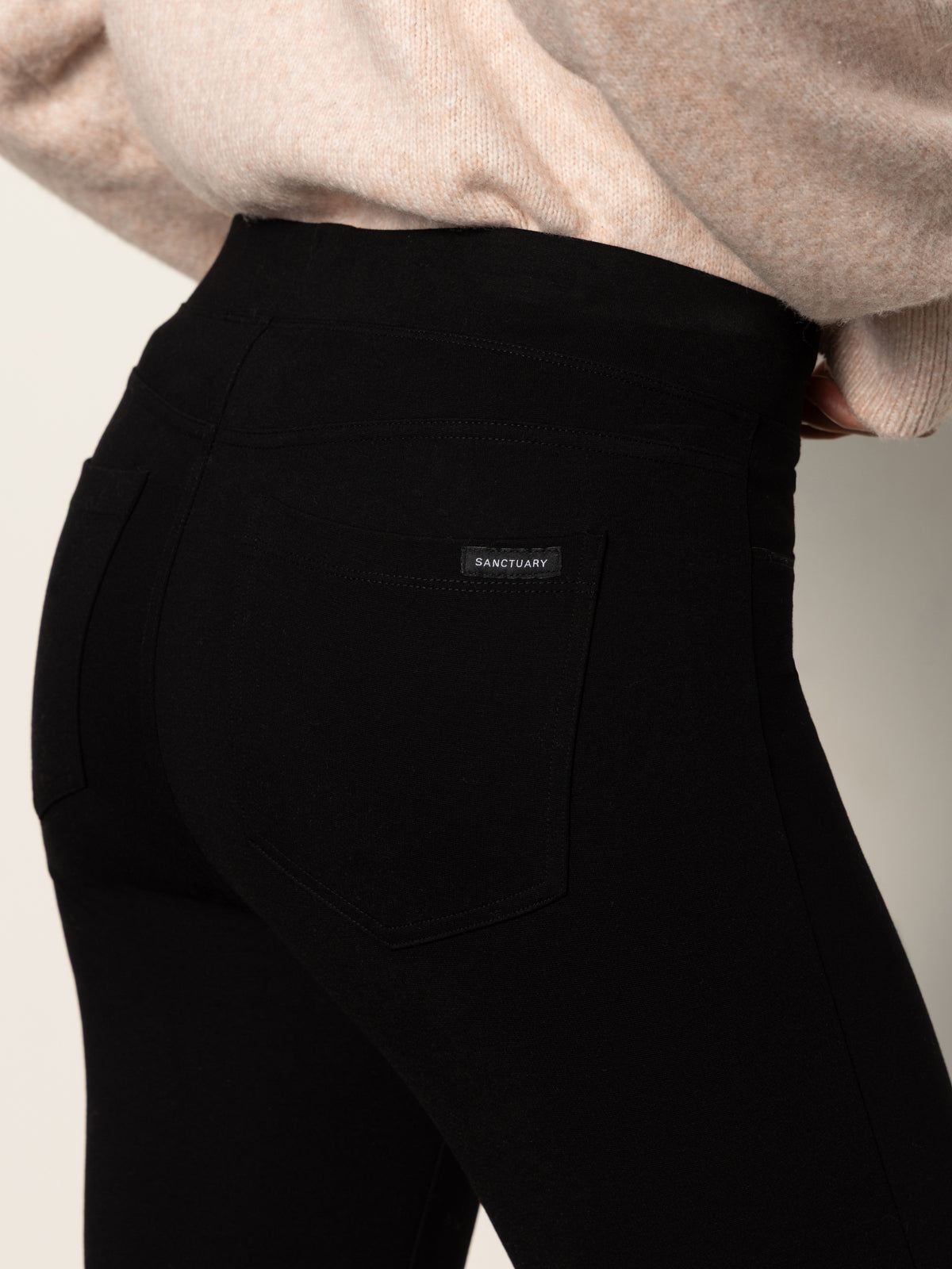 Close-up of a person wearing Sanctuary Clothing's 90s straight legging black pants and a light beige sweater. The pants feature a back pocket with a small "SANCTUARY" label; the person is turned slightly to the side.