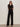 A woman stands against a plain background wearing Sanctuary Clothing's marine ponte pant in black, a sheer black long-sleeve blouse, heeled sandals, sunglasses, and minimal jewelry, arms crossed with a confident expression.