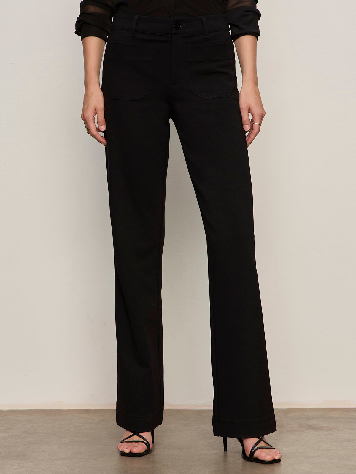 A person stands against a plain wall wearing Sanctuary Clothing’s marine ponte pant in black with black open-toe heels. Their arms are relaxed by their sides, and only their body from the shoulders down is visible.