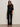A woman stands against a plain wall wearing black sunglasses, a sequined long-sleeve top, Sanctuary Clothing's velveteen brook bootcut pant in black petites with a belt, heeled sandals, and holding a black handbag by her side.