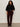A woman with curly hair stands against a plain light wall, wearing Sanctuary Clothing's velveteen brook bootcut pant in dark crimson (extended sizing), a black knit sweater, and black ankle boots, with one hand in her pocket and a neutral expression.