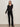 A woman with straight brown hair and a neutral expression stands against a plain background in black boots, a fitted long-sleeve lace top, and Sanctuary Clothing„¢s velveteen brook bootcut pant in black, accessorized with a belt.
