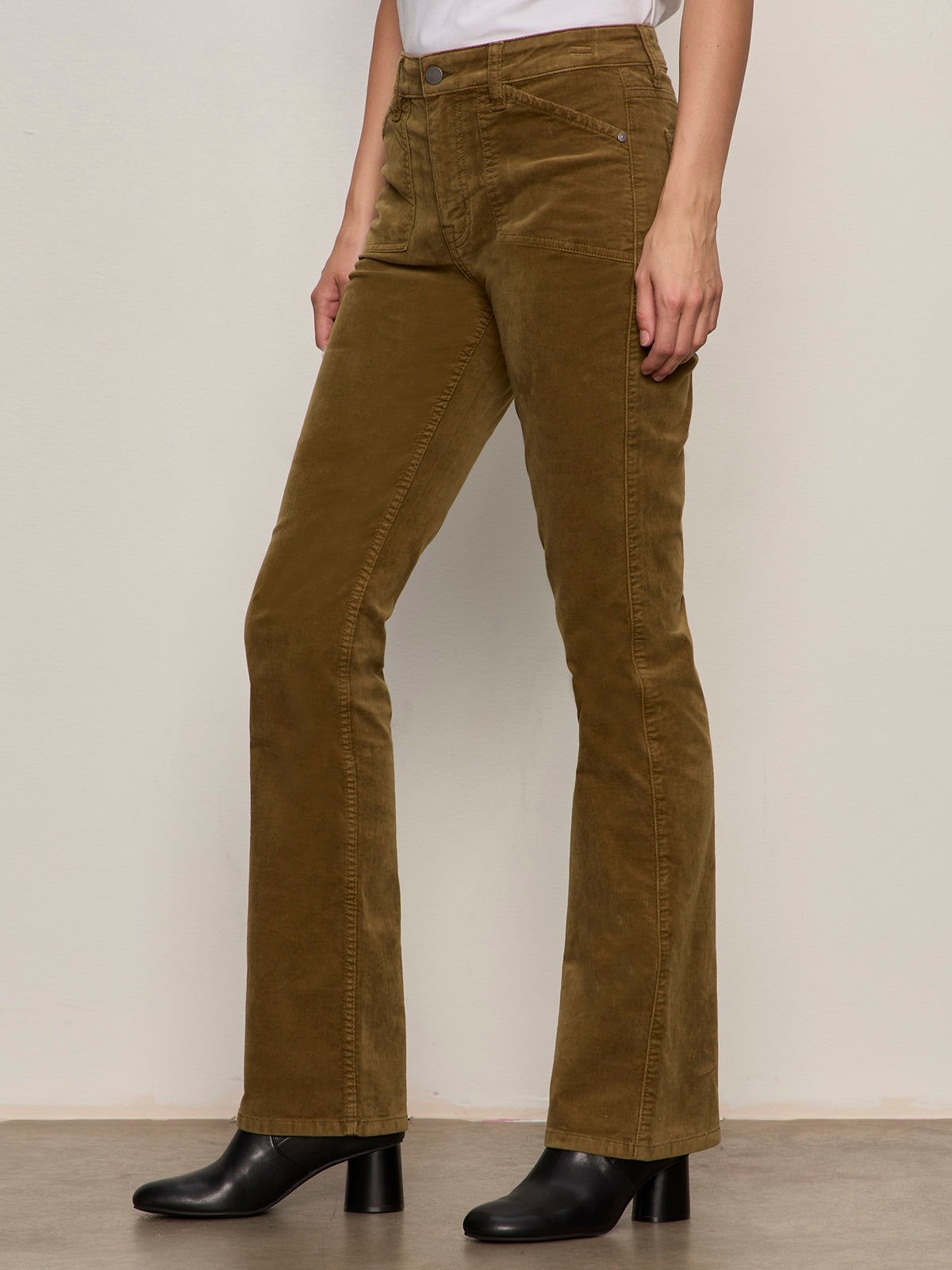 A person wears Sanctuary Clothing's velveteen brook bootcut pant in dirty martini, styled with a white top and black heeled ankle boots, standing against a light background.