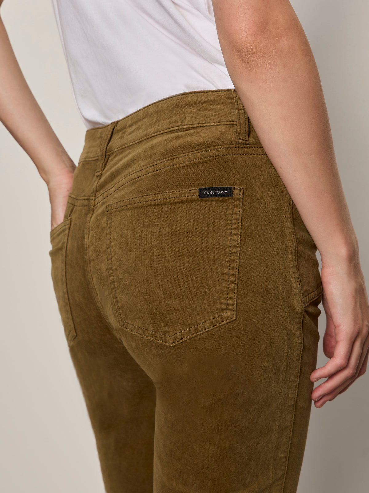 A person is shown from the back wearing Sanctuary Clothing's velveteen brook bootcut pant in dirty martini with a white shirt, one hand near the hip and the back pockets of the pants visible.