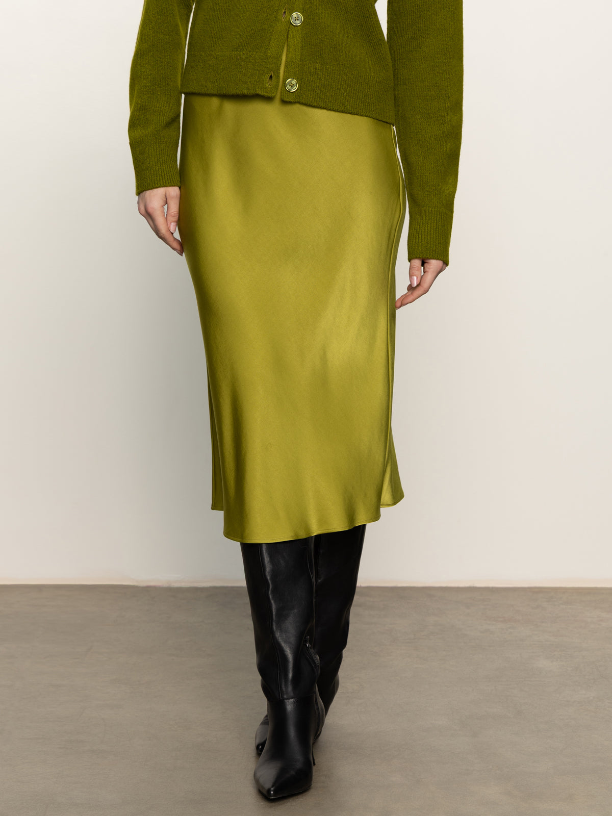 Only the lower half of a person wearing Sanctuary Clothing’s everyday midi skirt in martini olive is visible, paired with a green cardigan and black knee-high boots on a gray floor against a white wall.