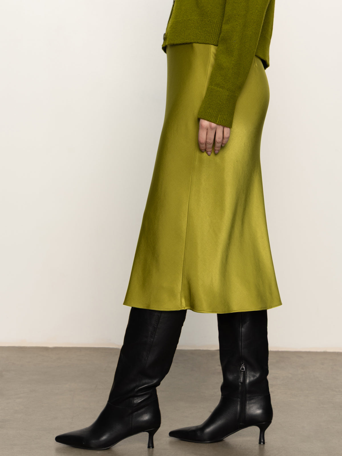 A woman stands indoors on a concrete floor against an off-white wall, wearing Sanctuary Clothing’s everyday midi skirt in martini olive, paired with a green sweater and black knee-high heeled boots.
