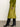 A woman stands indoors on a concrete floor against an off-white wall, wearing Sanctuary Clothing’s everyday midi skirt in martini olive, paired with a green sweater and black knee-high heeled boots.