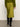 A person stands on a concrete floor facing away, dressed in a green sweater, black knee-high boots, and the Sanctuary Clothing everyday midi skirt in martini olive.