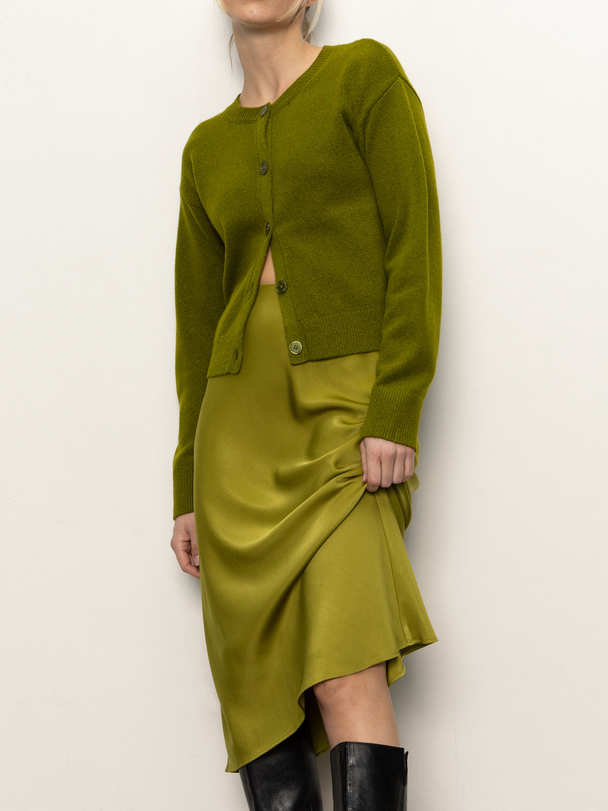 A person wearing a moss green cardigan and the Sanctuary Clothing everyday midi skirt in martini olive poses against a white background, partially lifting the skirt, with their face partly out of frame.