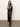 A woman stands with arms crossed, wearing Sanctuary Clothing's "everyday maxi skirt semi sweet" in dark brown satin, paired with a black and white patterned sweater and black heeled boots against a plain light background.