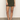 A person wears the Sanctuary Clothing marcela mini skirt kalamata, paired with a black top and black heeled sandals, standing on a gray floor against a plain white wall.