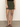 A person wears the Sanctuary Clothing marcela mini skirt kalamata, paired with a black top and black heeled sandals, standing on a gray floor against a plain white wall.