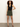 A woman with curly hair poses against a plain background in a sleeveless black button-up top, black flip-flops, and the Sanctuary Clothing marcela mini skirt in kalamata, with one hand on her hip and a neutral expression.