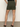 A person faces away, wearing the Sanctuary Clothing marcela mini skirt in kalamata with back flap pockets, paired with a black top and black heeled sandals, standing on a neutral floor against a plain wall.