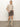 A woman stands indoors wearing the camden skirt french kitty by Sanctuary Clothing, a leopard print mini skirt, paired with a beige short-sleeve sweater and beige knee-high boots, hands in her pockets and head turned slightly to the side.