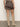 A person stands facing away, wearing the camden skirt french kitty by Sanctuary Clothing and knee-high beige suede boots with a low heel, against a plain light-colored wall and floor.