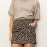 A person wears the Sanctuary Clothing camden skirt french kitty, a high-waisted leopard print mini skirt, with a short-sleeve beige sweater and one hand in the pocket, standing against a plain white background.