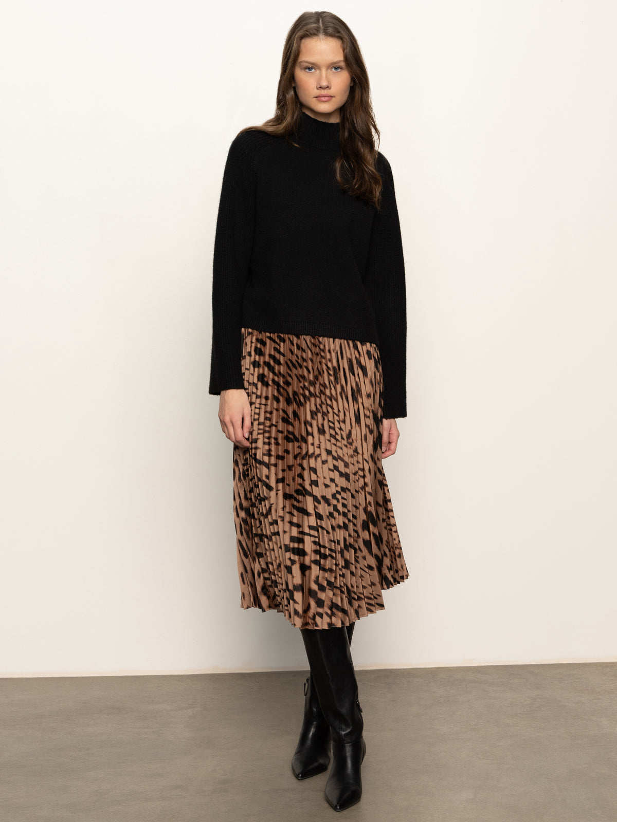 A woman stands against a white wall wearing the Sanctuary Clothing nostalgia pleated midi skirt in mocha leo, paired with a black long-sleeve top, black tights, and boots. Her hair is loose and she gazes at the camera with a neutral expression.