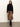 A woman stands against a white wall wearing the Sanctuary Clothing nostalgia pleated midi skirt in mocha leo, paired with a black long-sleeve top, black tights, and boots. Her hair is loose and she gazes at the camera with a neutral expression.