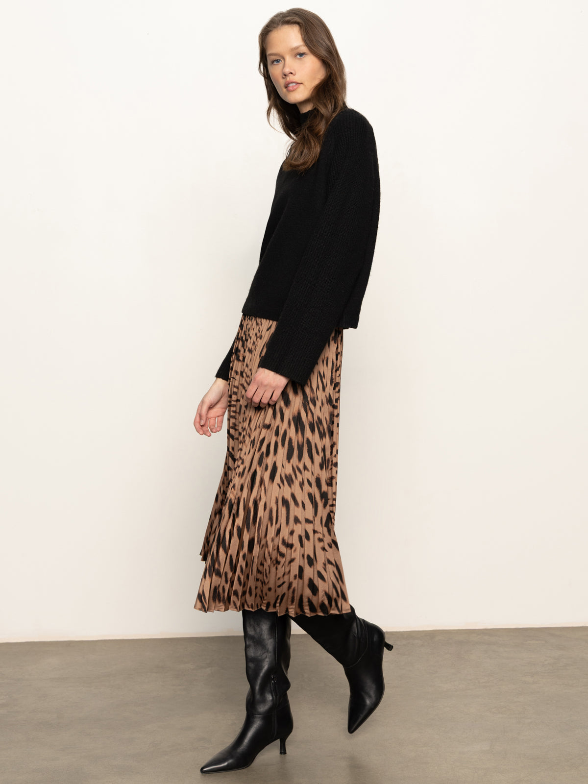 A woman stands against a plain background wearing the Sanctuary Clothing nostalgia pleated midi skirt in mocha leo, paired with a black sweater and knee-high black heeled boots, looking at the camera with a neutral expression.