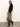 A woman stands against a plain background wearing the Sanctuary Clothing nostalgia pleated midi skirt in mocha leo, paired with a black sweater and knee-high black heeled boots, looking at the camera with a neutral expression.
