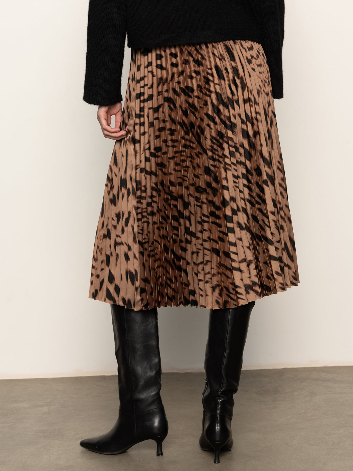 A person wearing the Sanctuary Clothing nostalgia pleated midi skirt in mocha leo, paired with knee-high black leather boots and a black top, stands on a gray floor against an off-white wall.